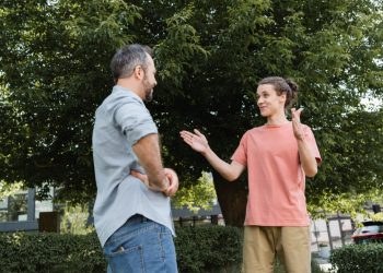happy teenage boy showing big gesture with hands near bearded father in park